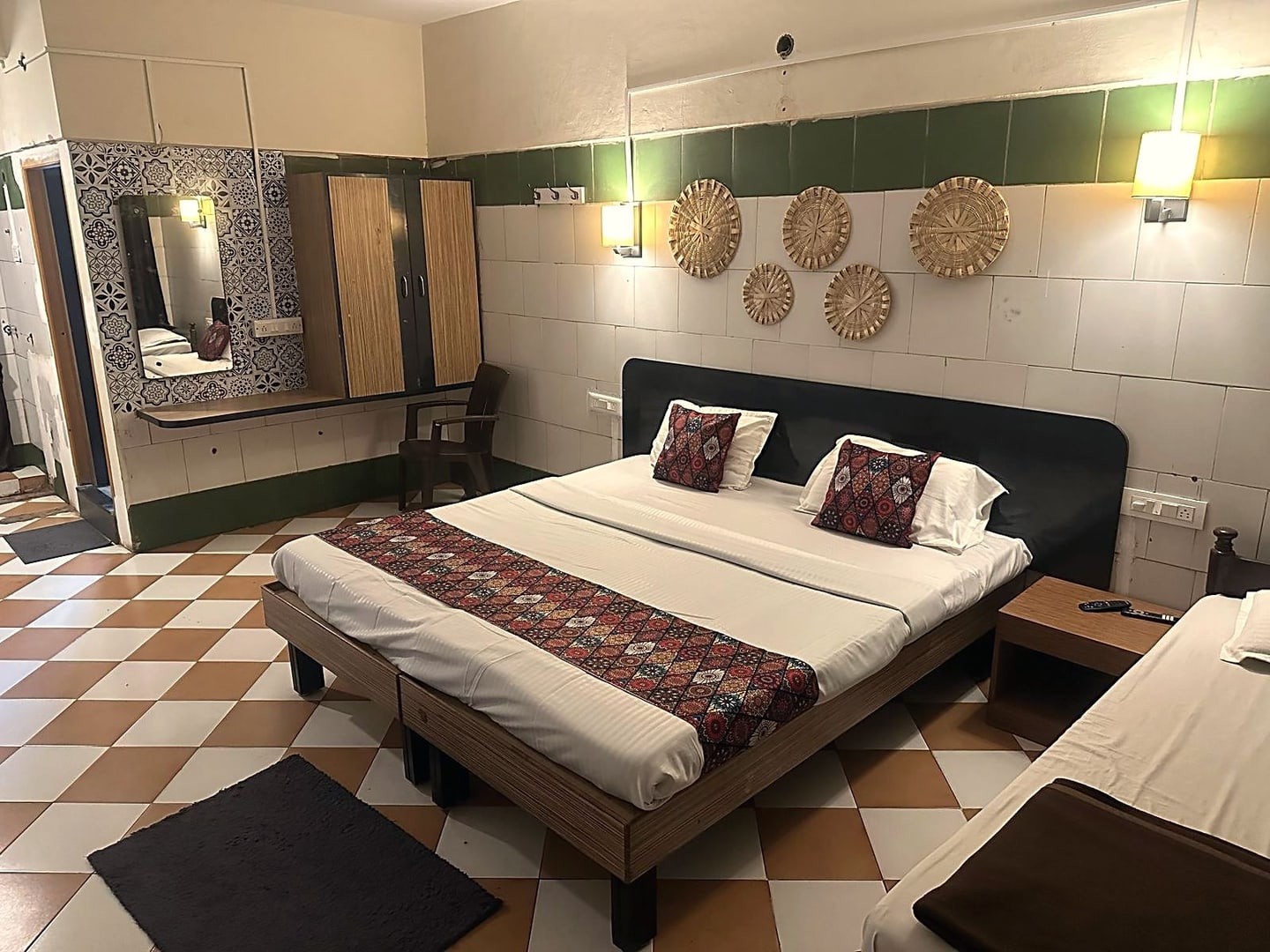 A Premium air conditioned room at Cibara Comforts in Harihar, with a made queen bed, deep green wainscot and cane wall discs lit by warm lamplight